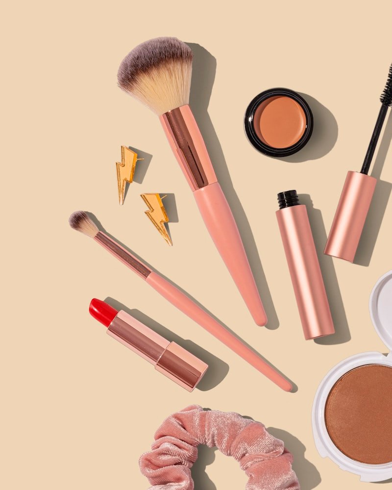 Beige flat lay of rose gold brushes, small gold earrings, a compact and a pink velvet scrunchie