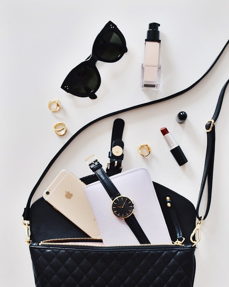Black crossbody bag laid flat with sunglasses, a watch, rings and a lipstick