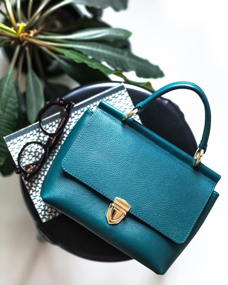 Teal green top-handle satchel with gold clasp beside a pair of sunglasses